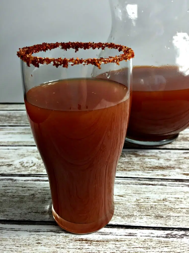 A glass of Dr Pepper with a spice-rimmed edge on a wooden surface, with a pitcher in the background.
