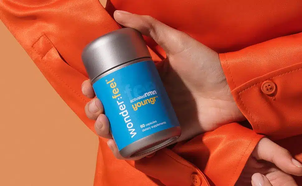 Person in an orange jacket holding a bottle of "wonderfeel" supplement, highlighting NMN Benefits, against an orange background.