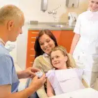 A dental professional shows a dental model to a young girl sitting in the dental chair with her mother beside her, focusing on oral health; a smiling dental assistant stands in the background.
