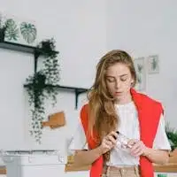 Serious female standing at table with used plastic bags and bottles near egg boxes and paper while taking off lid from glass jar sorting trash