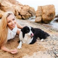 A woman with long blonde hair lies on the beach next to a black and white dog, enjoying a serene moment. Large rocks and the sea provide a picturesque backdrop, perfect for summer travel memories.