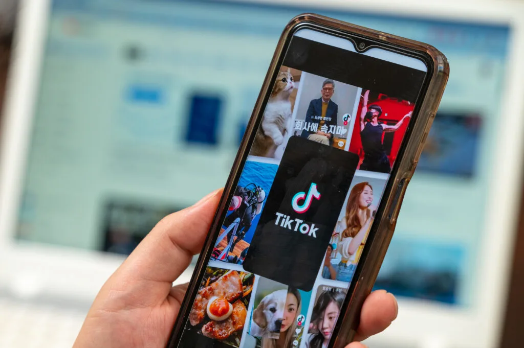 A hand holds a smartphone displaying the TikTok app with various video thumbnails, highlighting a growing number of TikTok followers, while a blurred computer screen features an online news site in the background.