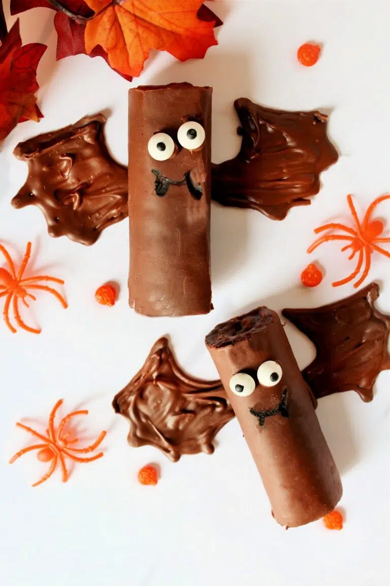 Two Halloween Bat Snack Cakes with chocolate rolls, adorned with candy eyes and bat wings made of chocolate, are surrounded by festive orange candy spiders and leaves.