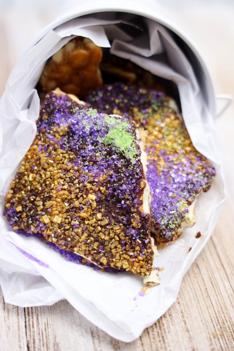 A metal container tipped over, spilling out Mardi Gras Cracker Candy, with pieces of bark-like treats coated in purple, green, and gold sugar crystals onto white paper—perfect for a sweet Fat Tuesday celebration.