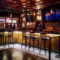 Dimly lit bar interior at The Ainsworth Hoboken with wooden stools lined up along the counter. Multiple screens are visible, and pendant lights hang from the ceiling, offering a cozy vibe typical of Hoboken restaurants.