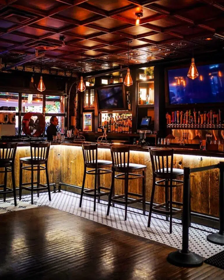 Dimly lit bar interior at The Ainsworth Hoboken with wooden stools lined up along the counter. Multiple screens are visible, and pendant lights hang from the ceiling, offering a cozy vibe typical of Hoboken restaurants.