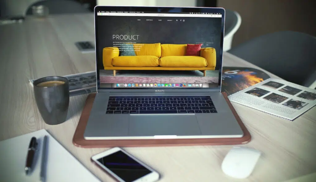 On a desk, an open laptop showcases an eCommerce site with a yellow sofa. Nearby, essentials like a smartphone, mug, open book, notebook, and wireless mouse enrich the workspace&mdash;a beginner's toolkit for navigating step-by-step guides to online shopping.