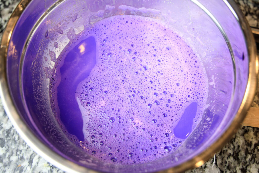 A stainless steel pot containing a frothy, purple liquid, perfect for whipping up those festive Mardi Gras Jello Shots, sits on a speckled countertop.