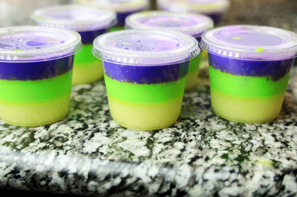 On a granite countertop sit small plastic cups filled with layers of purple, green, and beige gelatin, looking like festive Mardi Gras Jello Shots.