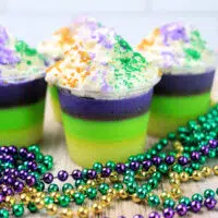 Four layered desserts in clear cups topped with whipped cream and colored sugar, reminiscent of Mardi Gras Jello Shots, placed on a wooden surface with purple, green, and gold beaded necklaces.