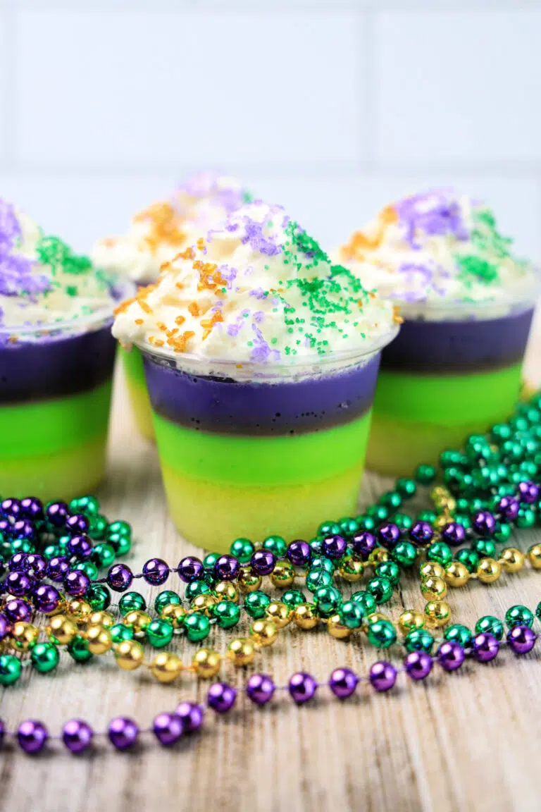 Four layered desserts in clear cups topped with whipped cream and colored sugar, reminiscent of Mardi Gras Jello Shots, placed on a wooden surface with purple, green, and gold beaded necklaces.