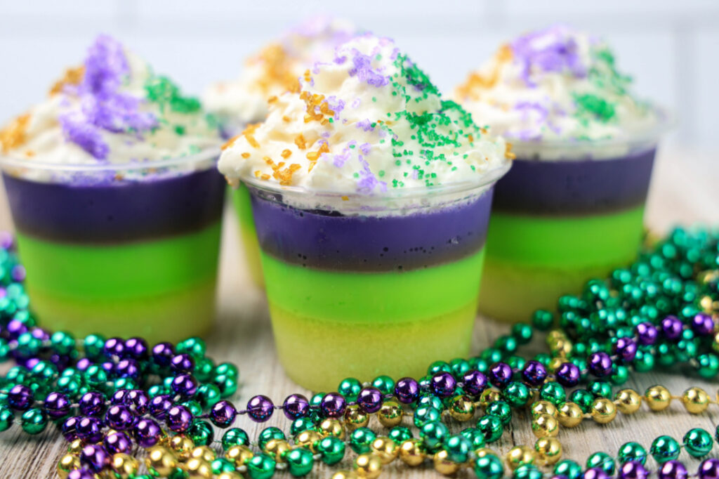Four layered desserts in clear cups with purple, green, and yellow layers mimic festive Mardi Gras jello shot recipes. Topped with whipped cream and colored sugar, these treats are perfect alongside vibrant beads in the foreground.