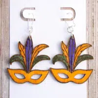 Wooden Mardi Gras Mask Earrings adorned with purple and green feathers gracefully presented on a pristine white display card.