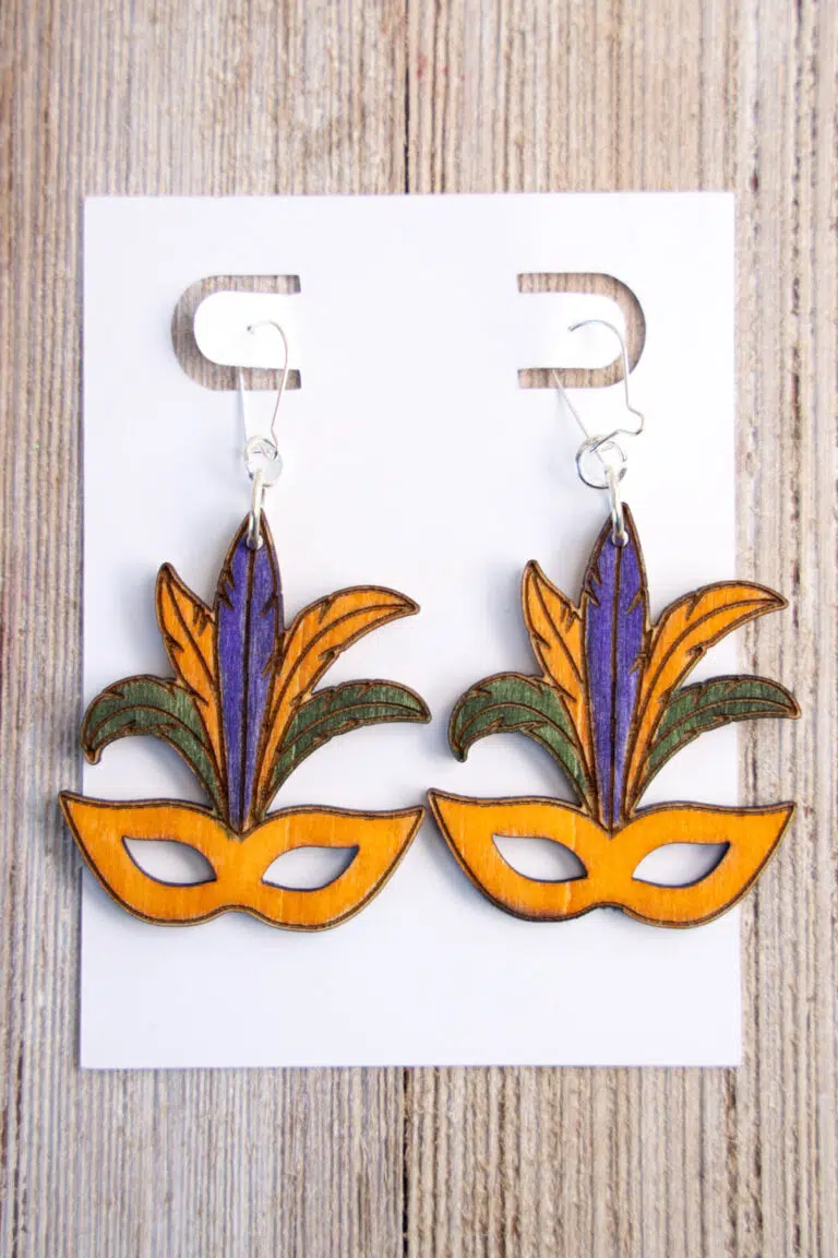 Wooden Mardi Gras Mask Earrings adorned with purple and green feathers gracefully presented on a pristine white display card.