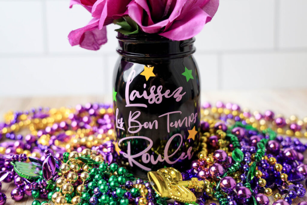 Purple mason jar with "Laissez les bon temps rouler" surrounded by colorful Mardi Gras beads and a decorative crown.