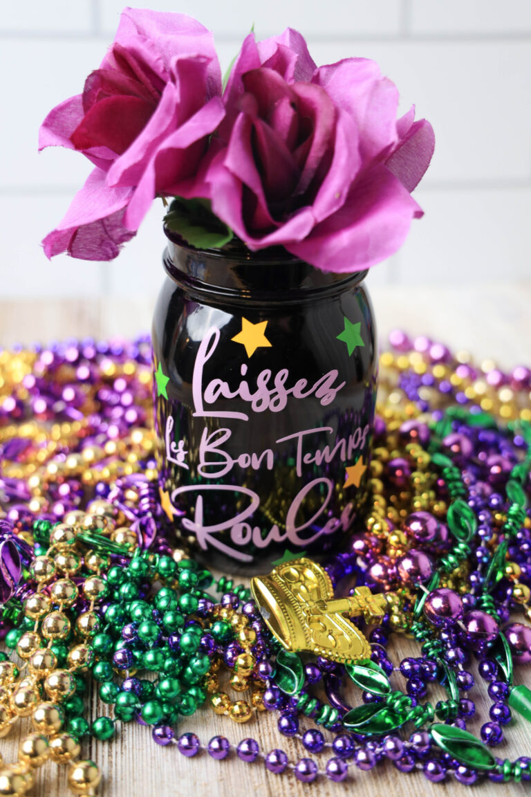 A black Mason jar adorned with the phrase "Laissez les bon temps rouler" is encircled by purple, green, and gold beads alongside pink flowers, resting on a wooden surface. Perfect for adding a touch of Mardi Gras spirit to your decor or following along with a creative tutorial.