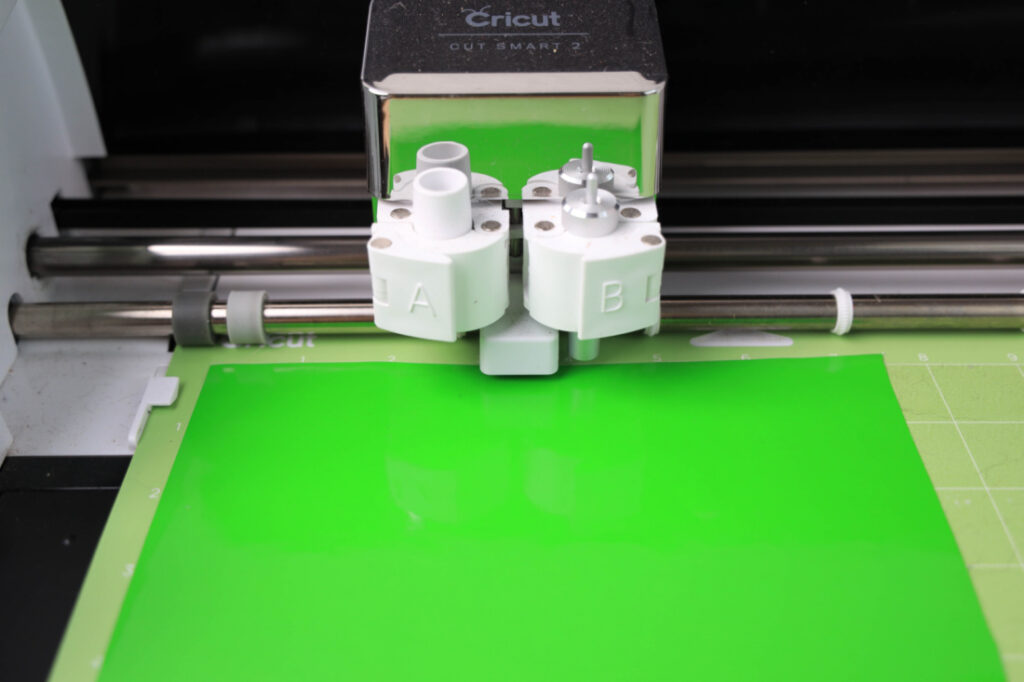 A Cricut machine, resembling a Mardi Gras celebration with its vibrant energy, cuts a bright green sheet on a mat. Two tool holders labeled A and B stand ready, like dedicated instructors in a crafting tutorial.