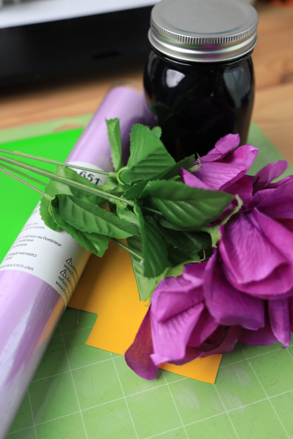 A tutorial on crafting Mardi Gras decorations: artificial purple flowers and a roll of purple paper lay neatly on a green cutting mat, next to a Mason jar filled with dark liquid.