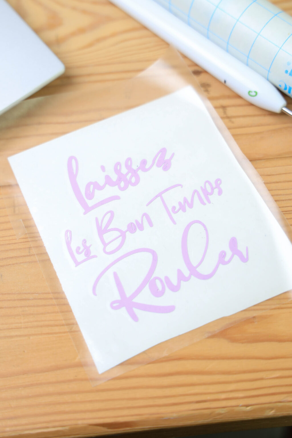 A clear sheet on a wooden surface displays the phrase "Laissez Les Bon Temps Rouler" in pink script, evoking the vibrant spirit of Mardi Gras.
