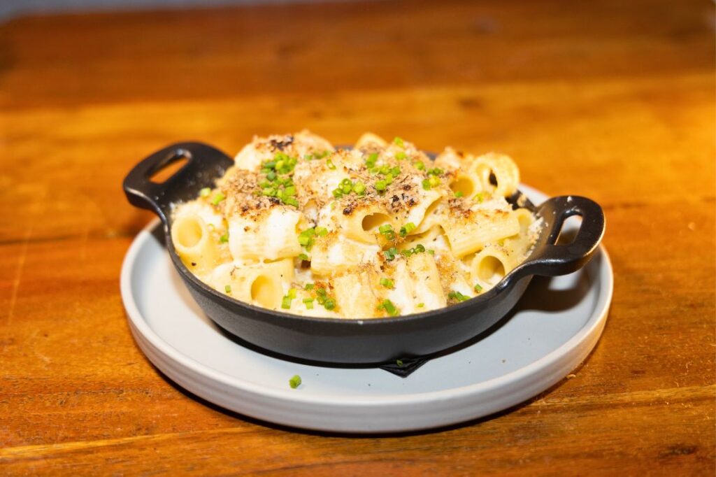 A dish of baked rigatoni pasta with melted cheese and chopped chives, served in a black cast iron pan on a white plate, reminiscent of The Ainsworth Hoboken's style, placed on a wooden table.