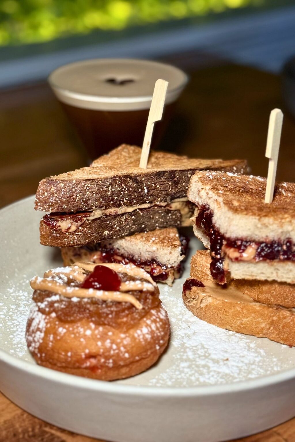 A plate features a peanut butter and jelly sandwich, a donut topped with the classic combo, and a coffee nestled behind. This delightful spread showcases the creativity found in Hoboken restaurants like The Ainsworth Hoboken.