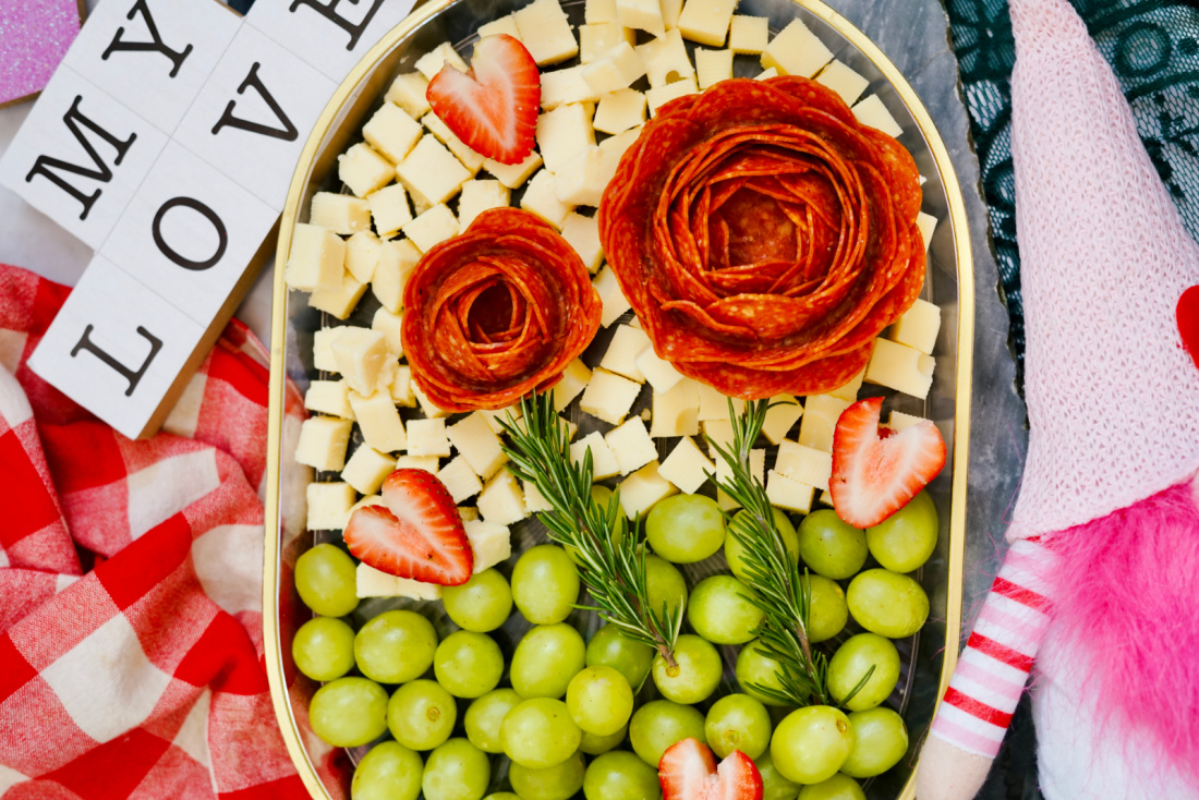 A Valentine's Day charcuterie board featuring a platter with green grapes, cubed cheese, pepperoni roses, strawberry slices, and rosemary. Nearby are printed tiles spelling "MY LOVE" and a pink hat with a white pom-pom.