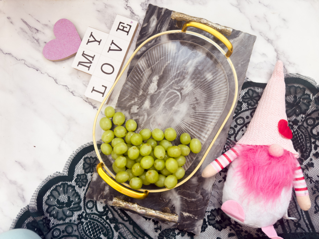 A delightful Valentine’s Day charcuterie board features a tray with green grapes, a pink gnome figurine, and letter tiles spelling "MY LOVE" on a marble surface with lace lining.