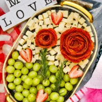 A decorative Valentine's Day charcuterie board with cheese cubes, salami roses, strawberries, grapes, and rosemary. A sign reads 