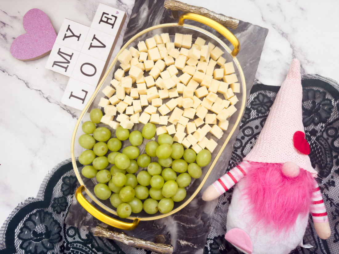 A Valentine’s Day charcuterie board showcases cheese cubes and green grapes on a marble surface, accompanied by a pink gnome doll and "MY LOVE" letter blocks.