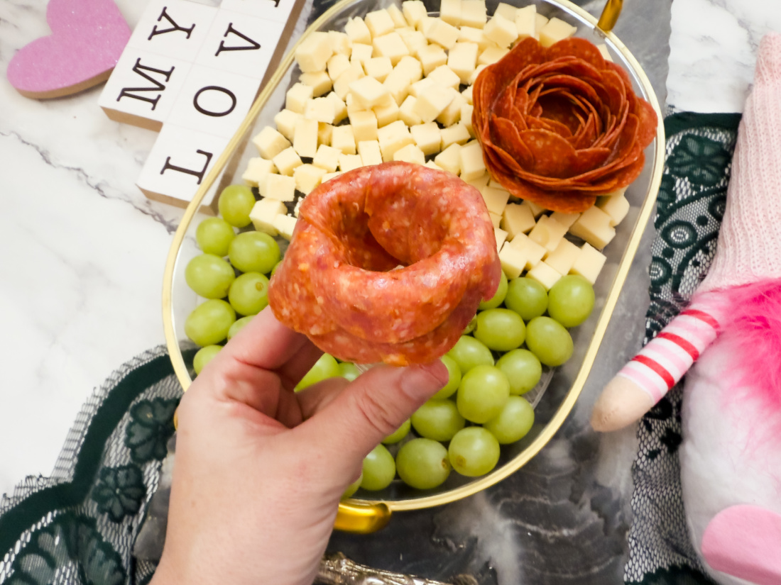 A hand holds a salami slice shaped into a cup over a plate adorned with cubed cheese, green grapes, and floral-shaped salami—a perfect creation for Valentine's Day charcuterie boards. In the background, blocks spell out "My Love," enhancing the festive mood.