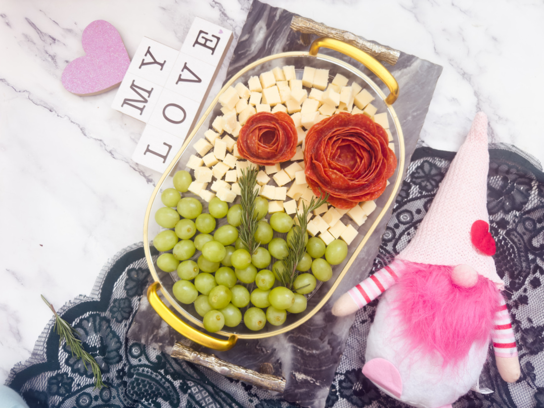 A Valentine's Day charcuterie board featuring cheese cubes, green grapes, and pepperoni roses. Nearby, a plush gnome, "MY LOVE" tiles, a pink heart, and lace fabric add a touch of romance.