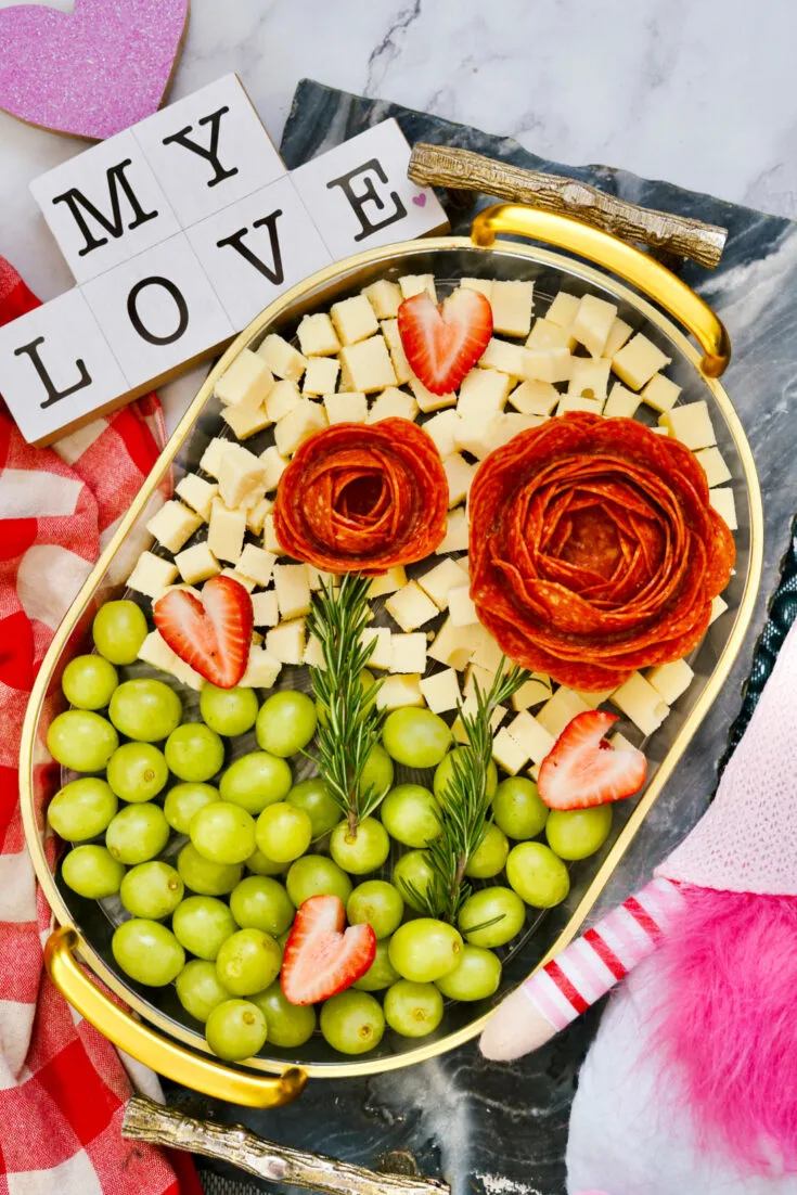 A decorative Valentine's Day charcuterie board with cheese cubes, salami roses, strawberries, grapes, and rosemary. A sign reads 