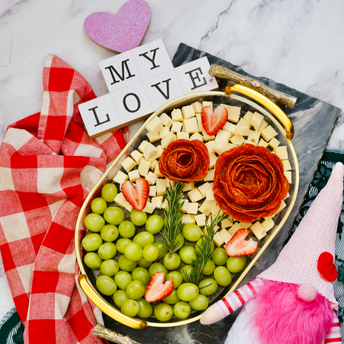 A delightful Valentine's Day charcuterie board features an oval platter with grapes, cheese cubes, strawberries, and salami roses. Accompanied by a gnome figurine, a pink heart, and "MY LOVE" spelled out with tiles on the marble surface for added charm.