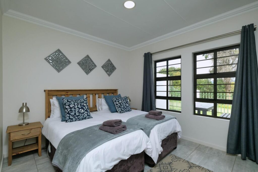 Bedroom with two beds, gray blankets, and decorative pillows. Wall art above bed, nightstand with lamp, large window with gray curtains, and beige walls create a perfect home style vibe. Ideal for those looking to achieve an effortless room revamp.