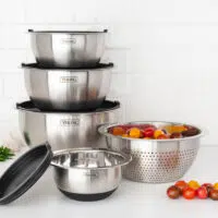 A set of four stacked stainless steel Viking mixing bowls, one with a lid—a must-have among kitchen essentials—and a colander filled with tomatoes on a white kitchen counter.