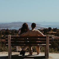 Two people sit on a bench overlooking a cityscape and coastline on a clear day.