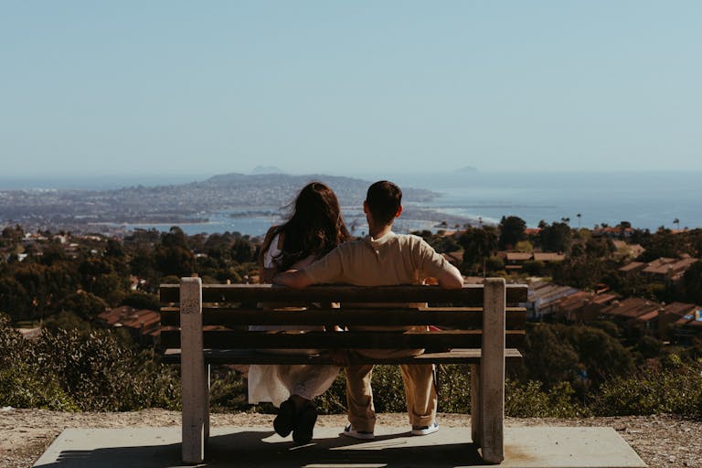 Two people sit on a bench overlooking a cityscape and coastline on a clear day.