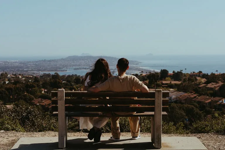 Two people sit on a bench overlooking a cityscape and coastline on a clear day.