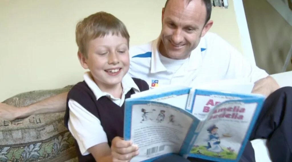 An adult and a child sit on a couch, smiling and reading a book together, highlighting the importance of early diagnosis in supporting children’s development.