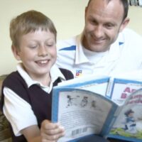 An adult and a child sit on a couch, smiling and reading a book together, highlighting the importance of early diagnosis in supporting children’s development.