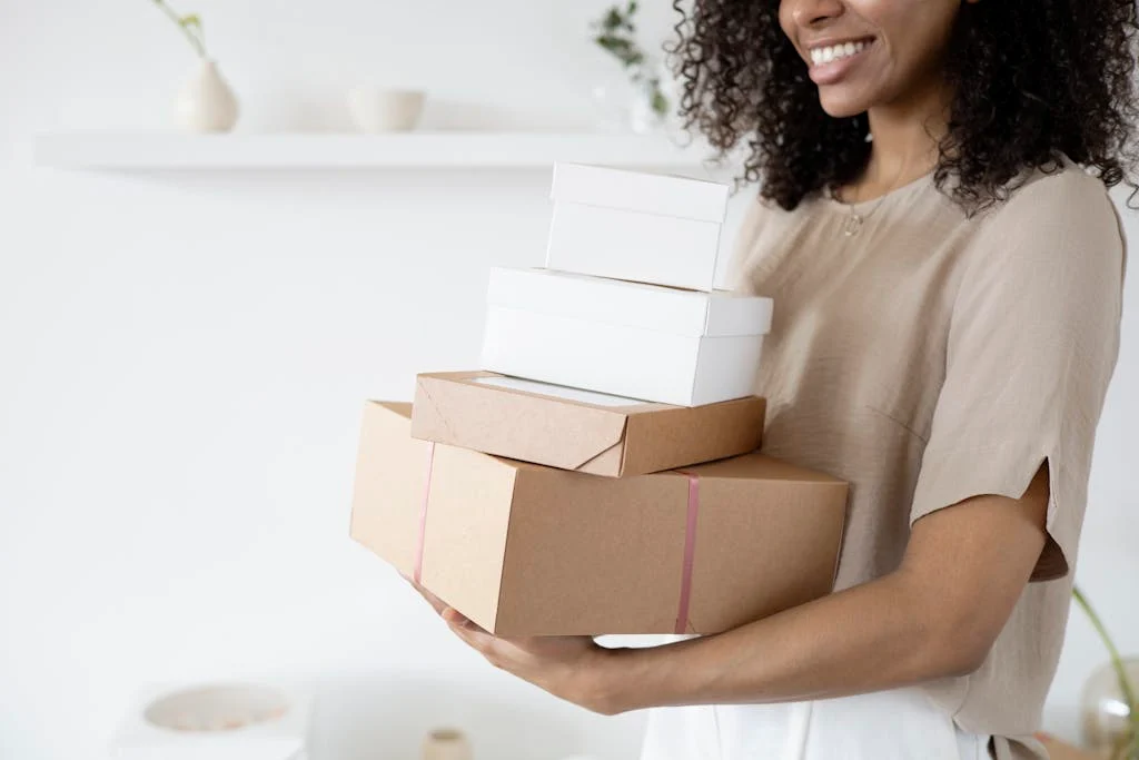 Woman with curly hair carrying stack of packaging boxes in a bright room.
